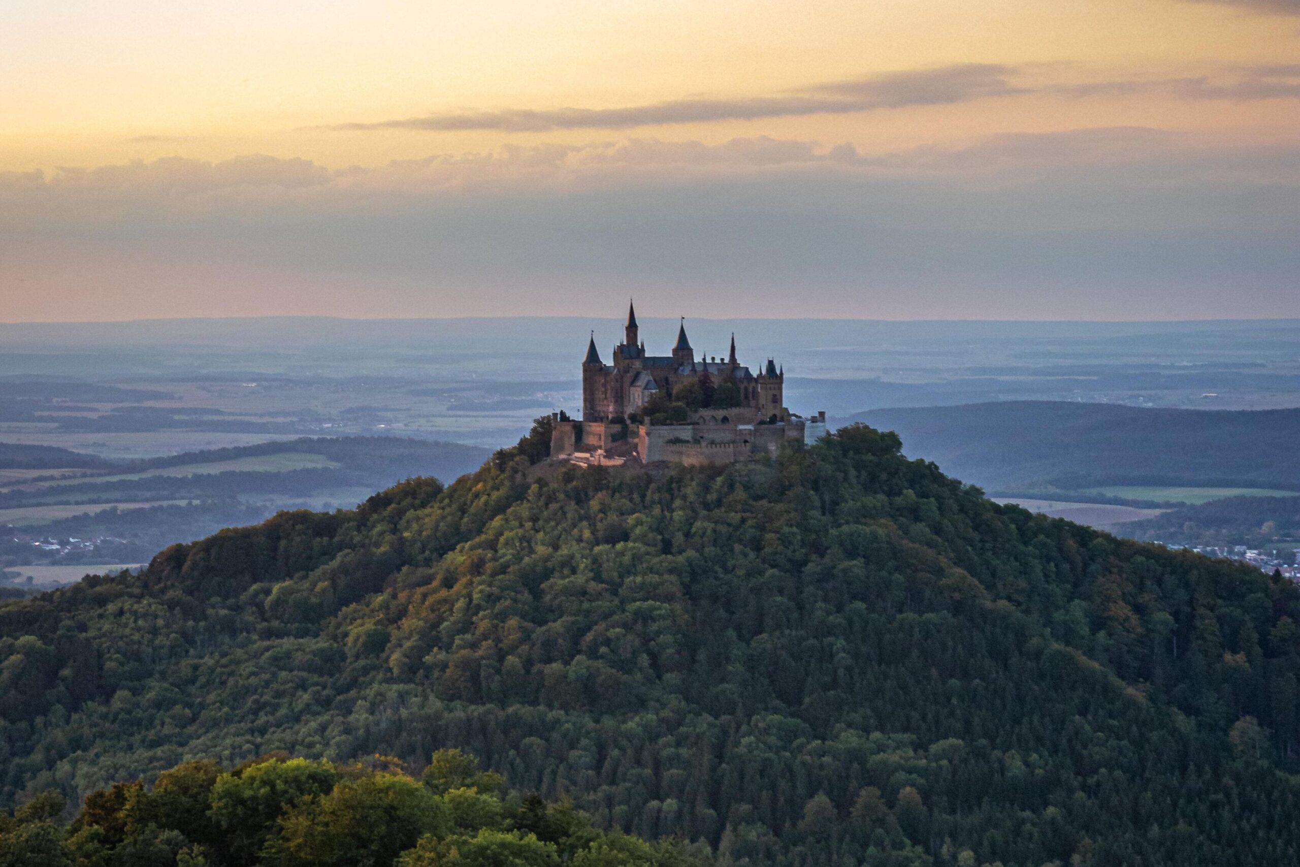 Weitwinkelaufnahme einer prachtvollen, neugotischen Burg mit vielen Türmen, die isoliert auf der bewaldeten Kuppe eines steilen Berges thront, im Hintergrund das weite, flache Albvorland im Abendlicht.