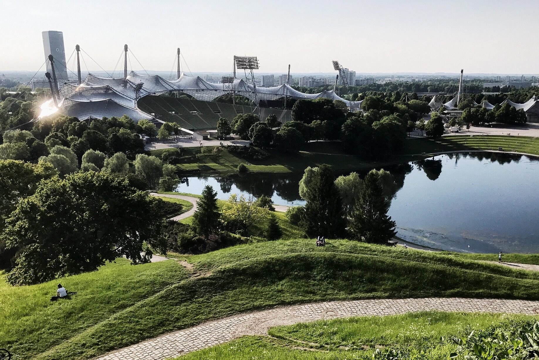Weitblick vom Olympiaberg auf den Olympiasee und das markante Stadion mit seiner transparenten Zeltdach-Architektur, eingebettet in eine hügelige Parklandschaft in München, mit dem O2-Hochhaus im Hintergrund.