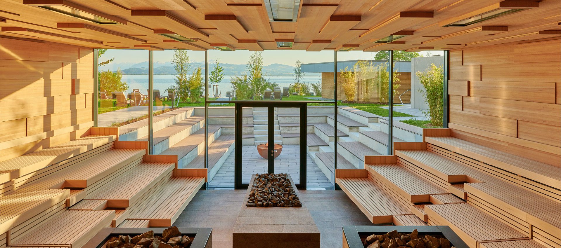 Großzügige Theatersauna der Therme Lindau mit mehrstufigen Holzliegen, dekorativem Saunaofen und bodentiefen Fensterfronten mit Blick auf den Bodensee und die Alpen